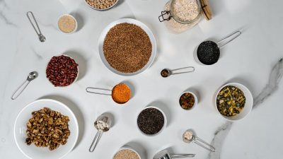 Flat lay of various dried grains, seeds, and beans arranged in bowls and measuring cups on marble.